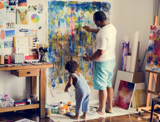 Black artist dad doing his art work with his child sitting nearby