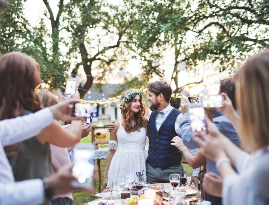 Guests with smartphones taking photo of bride and groom at wedding reception outside.